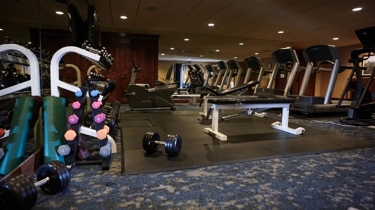 This image features a well-equipped interior fitness space, with a set of dumbbells, treadmills, and a weight bench visible. The clean, bright environment is designed for active pursuits and group activities, emphasizing communal recreation.
