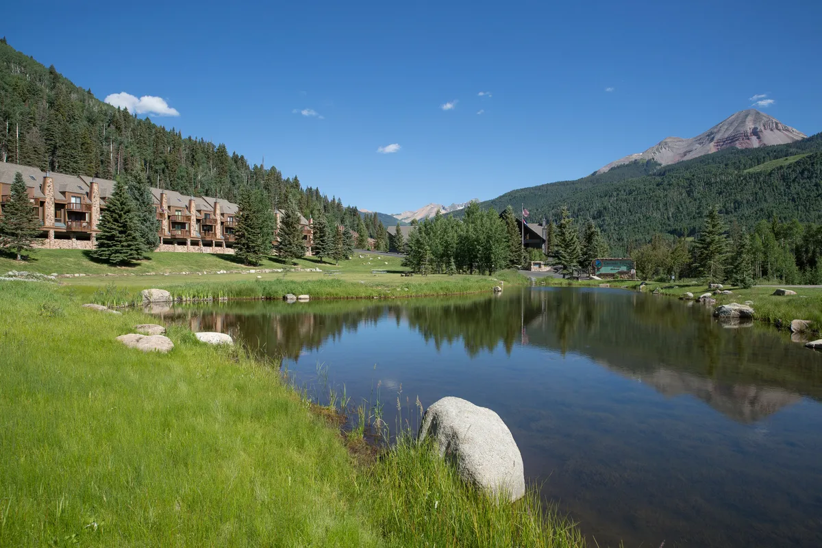 A serene lake lies in the foreground, edged by wooden lodge-style buildings and enveloped by lush greenery and mountains. The peaceful scene hints at family-friendly activities and relaxation amidst nature, extending the experience beyond winter adventures.