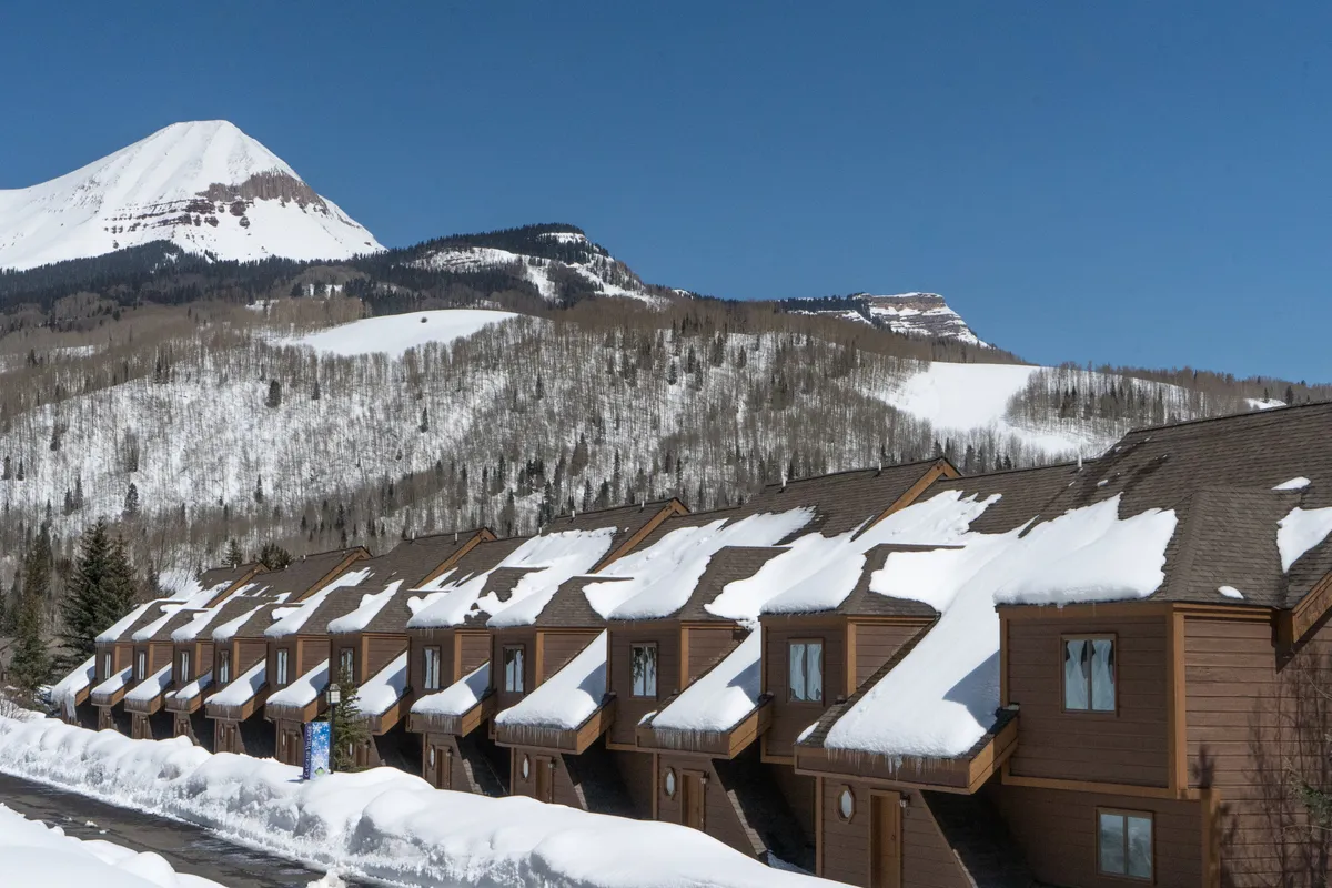 A tranquil row of chalets sits under a blanket of snow, with dramatic mountains creating a majestic backdrop. The scene suggests warmth and comfort after a day outdoors, inviting families to imagine cozy evenings and unforgettable winter days.