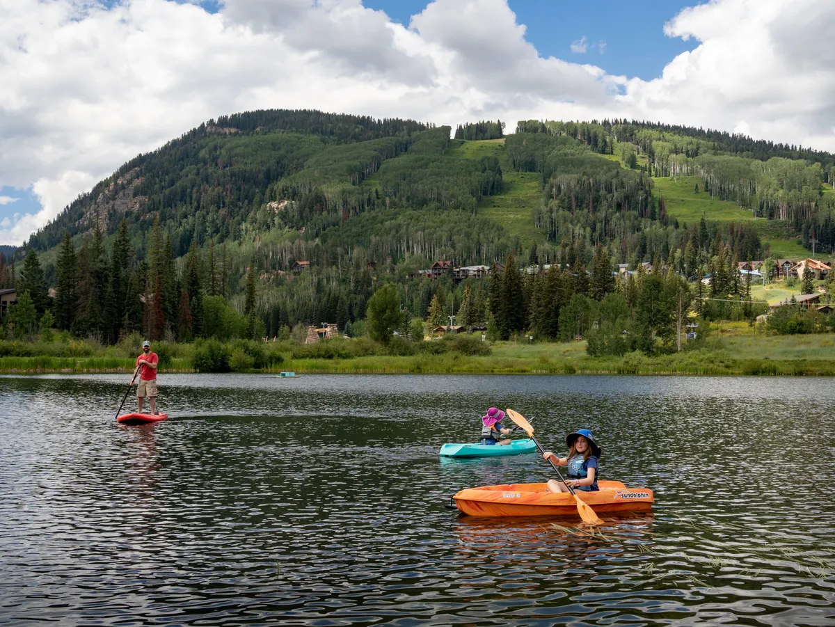 This image captures kayakers and a paddleboarder enjoying a sunlit lake with majestic mountain peaks in the backdrop. The water reflects both the paddlers and the surrounding landscape, evoking a sense of adventure and connection to the outdoors.