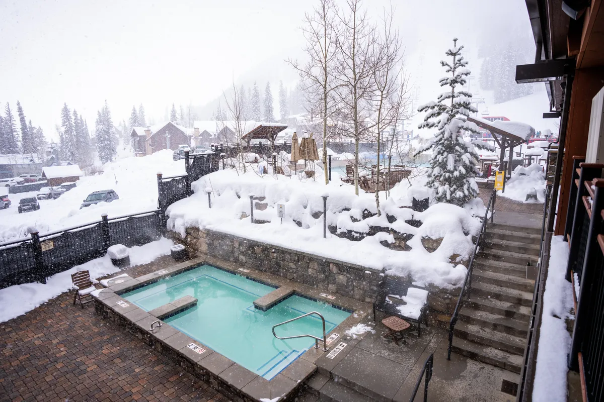A steaming outdoor hot tub nestled among snow-covered trees and a rustic stone wall. The inviting water contrasts beautifully with the snowy landscape, evoking a sense of cozy relaxation and luxurious escape even in colder months.