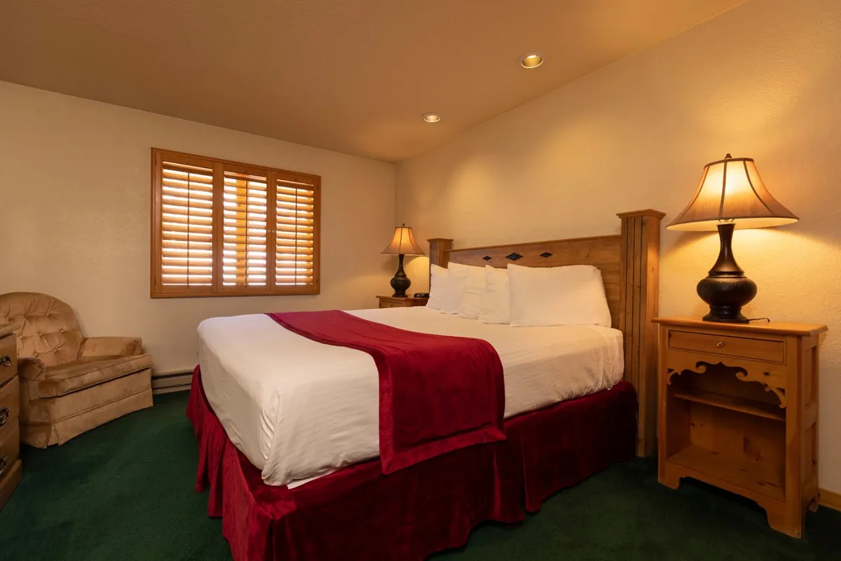 This image showcases a cozy bedroom featuring a bed with plush linens, a warm wooden headboard, and inviting lamps on nightstands. A window with shutters provides a glimpse of the tranquil mountain setting, and the overall mood is comfortable and restful.
