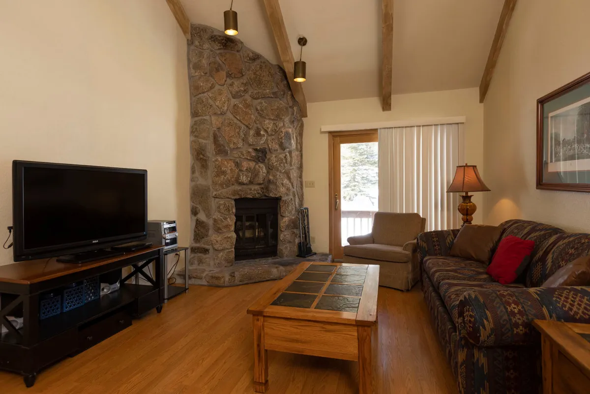 The image showcases a cozy living room with a stunning stone fireplace as the centerpiece, a plush sofa adorned with inviting cushions, a rustic wooden coffee table, and a large television—creating a perfect setting for relaxation and togetherness after a day in the mountains.