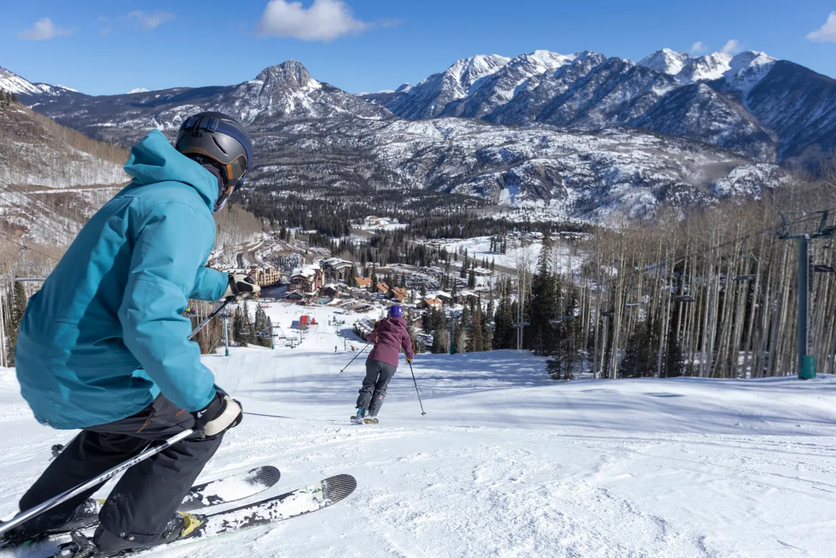 This image features skiers descending pristine, snow-covered slopes with sweeping views of the surrounding mountains. At the base lies a cozy resort village nestled in the valley, hinting at both adventure and relaxation. The scene vibrantly captures the thrill of winter sports and the inviting atmosphere of a mountain resort.