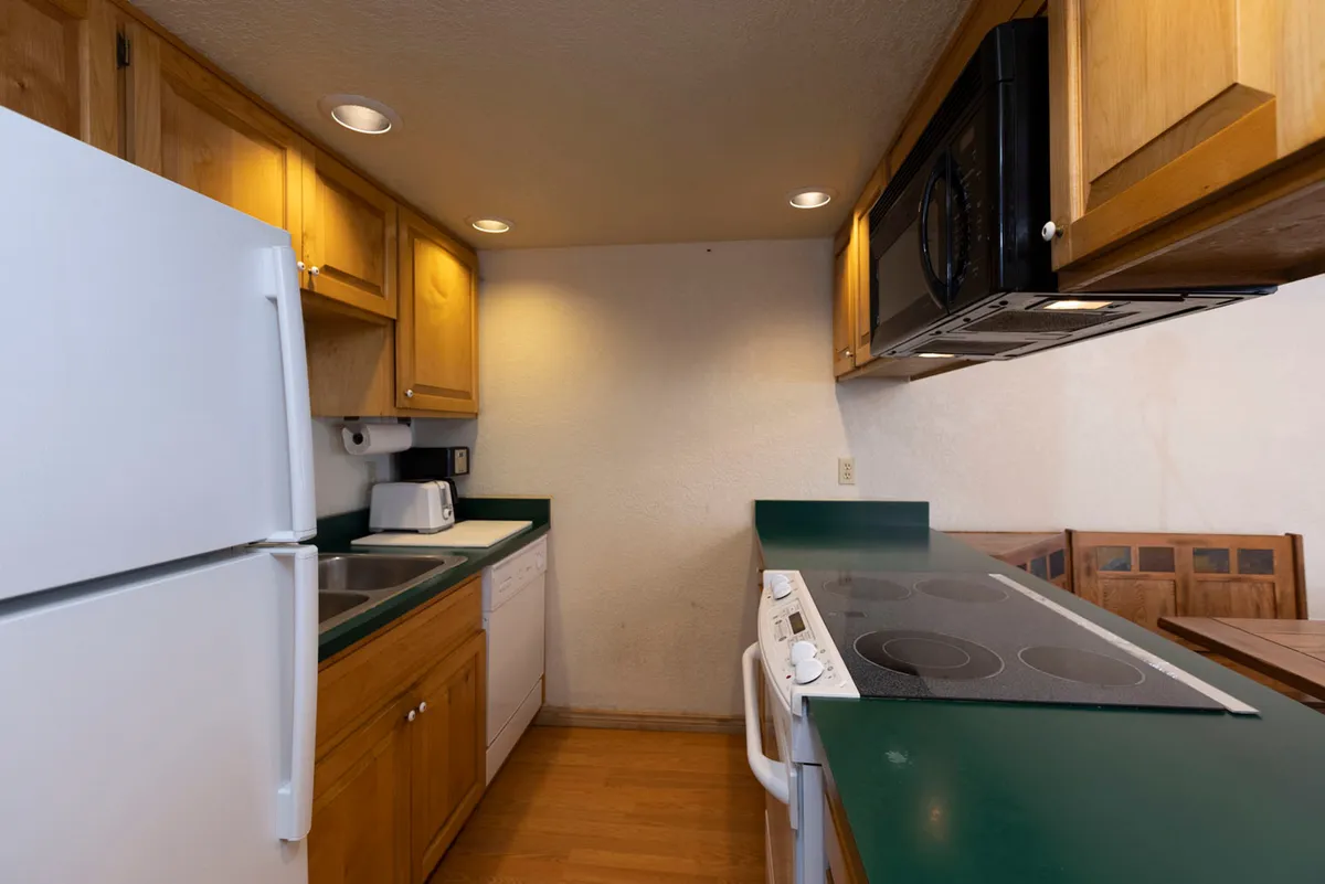 A broad perspective that incorporates kitchen appliances, wooden cabinets, a green countertop, and the dining area in the background. The atmosphere is inviting and practical, suggesting space for meal prep, entertaining guests, and sharing stories over dinner. The visual connection between the kitchen and dining area reinforces the open-concept design.